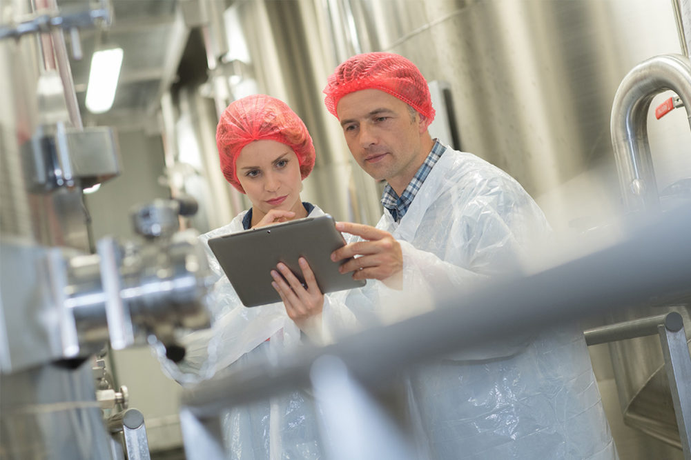 Two employees in red hair nets discussing over clipboard (©STOCKR - STOCK.ADOBE.COM)